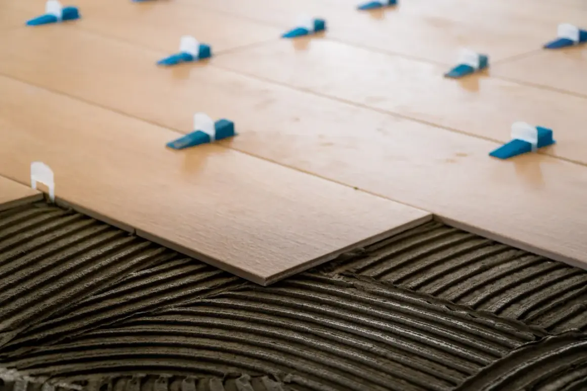 Pose de carrelage en quinconce sur lit de mortier. Des cales bleues et blanches assurent l'espacement régulier des lames imitation bois.