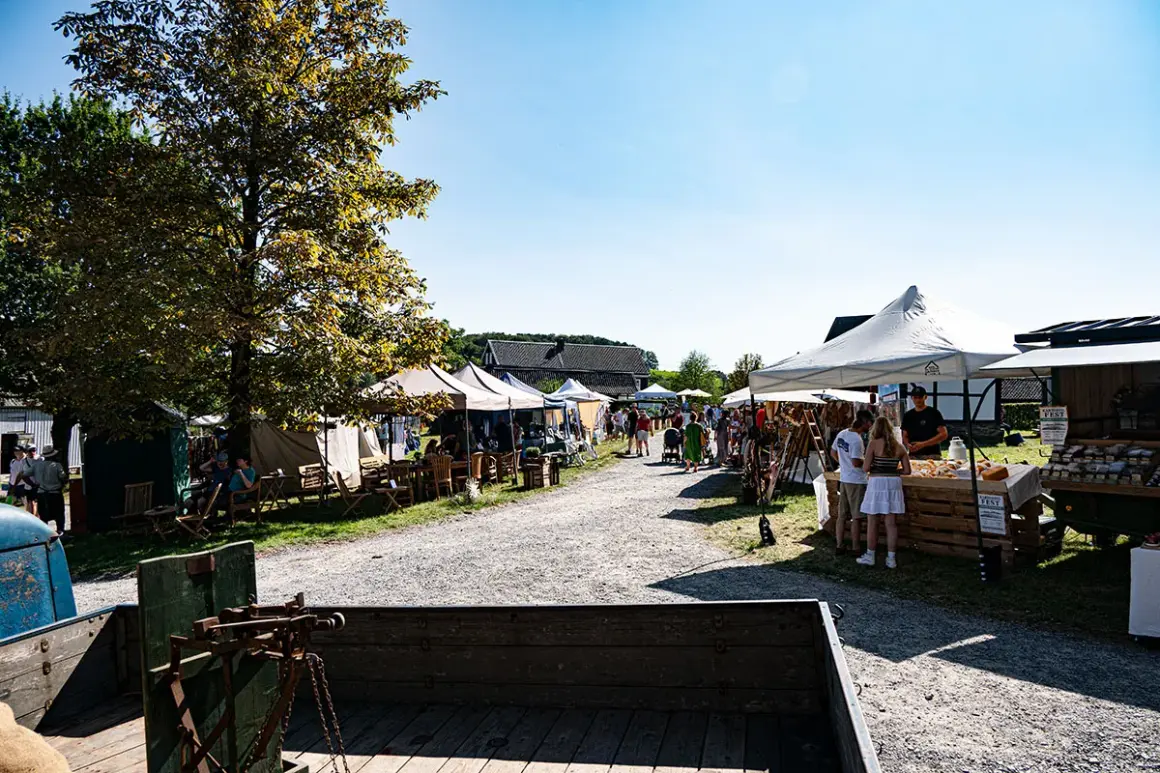 Besucher bummeln über den Bauernmarkt Lindlar, vorbei an Ständen mit Waren und Sitzgelegenheiten unter Bäumen.