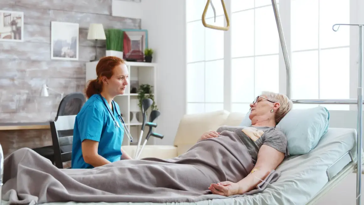 Pielęgniarka rozmawia z pacjentką leżącą w łóżku. W tle widać kule, co sugeruje, że pacjentka może potrzebować wsparcia w odżywianiu dojelitowym.