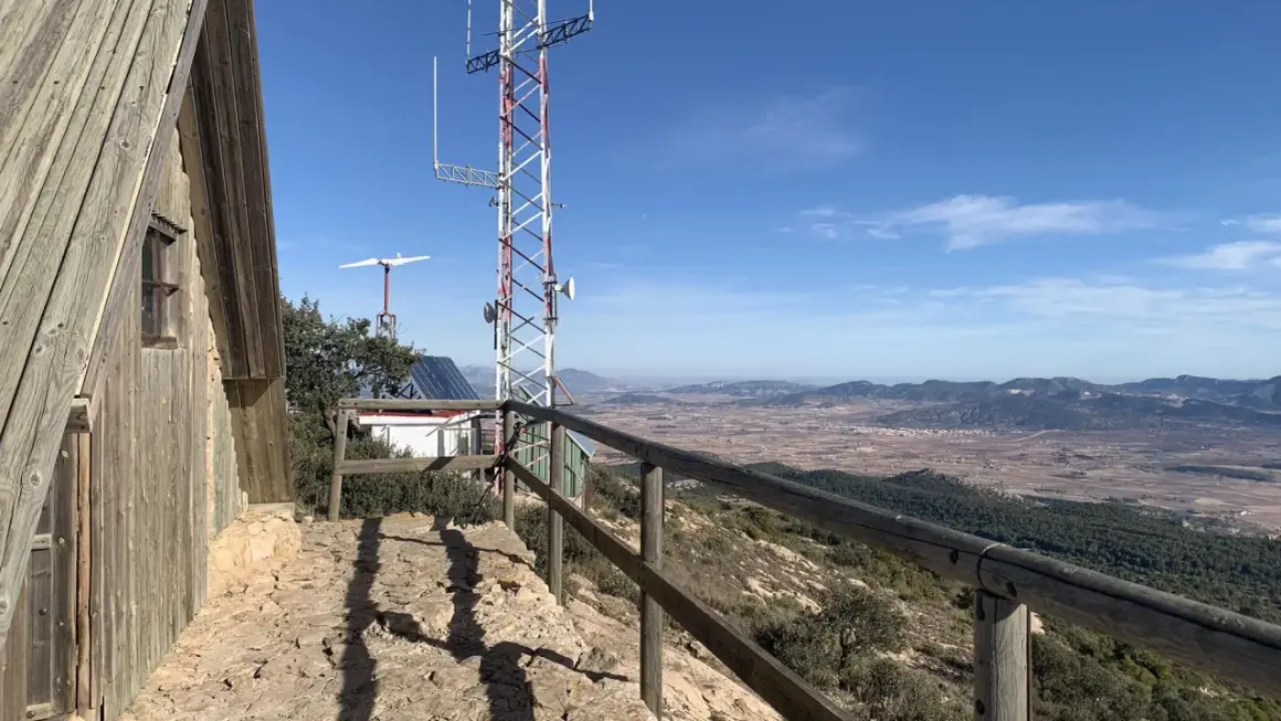 Vista panorámica desde la sierra del Carche, con una cabaña de madera, antena de comunicaciones y un paisaje de campos y montañas.