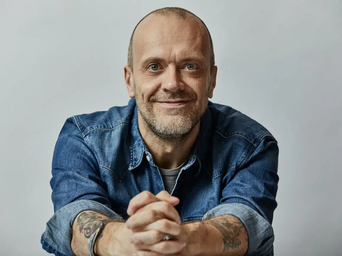 Uomo con barba corta, occhi azzurri, indossa una camicia di jeans. Le sue mani sono intrecciate, eccoti testo.