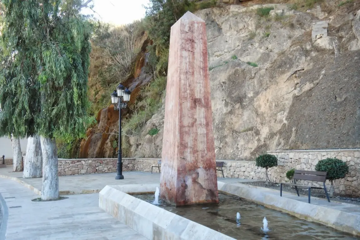 Obelisco en fuente con cascada alhama de almeria al fondo. Árboles y bancos invitan al descanso.