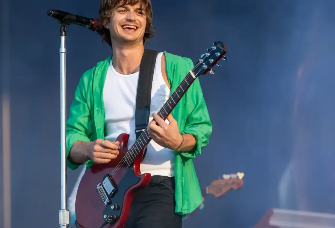 Joe Keery w zielonej koszuli gra na czerwonej gitarze elektrycznej na scenie.