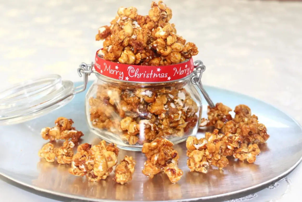 A jar overflowing with caramel popcorn, tied with a red "Merry Christmas" ribbon, sits on a silver platter with scattered popcorn.