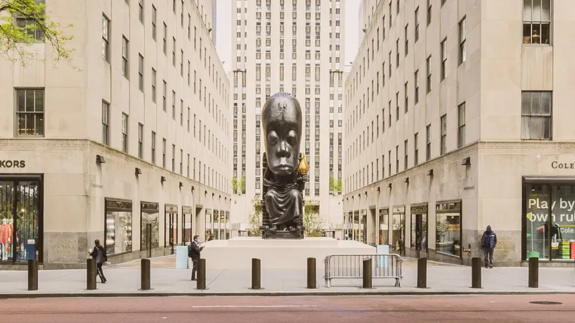 A monumental sculpture of a seated figure with a large head, holding a torch, stands as a striking example of public art between two imposing buildings.