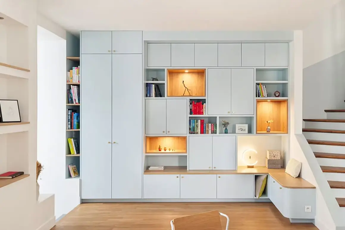 Un mur habillé de rangements modulables bleu pâle, avec des niches éclairées et une banquette intégrée près d'un escalier en bois.