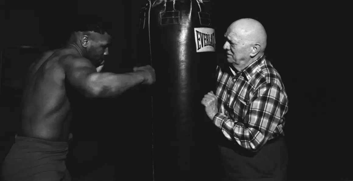 Mike Tyson trains with his manager, a bald man in a plaid shirt, in a dimly lit gym.