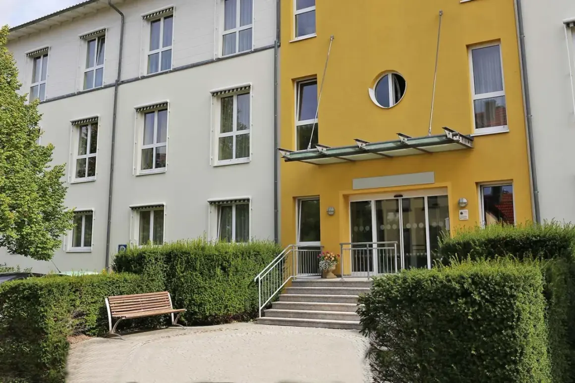 Gelbes Gebäude mit runden Fenster, Eingang mit Glasdach und Stufen. Ein Ort, der §43b SGB XI-Leistungen bietet, lädt zum Verweilen ein.