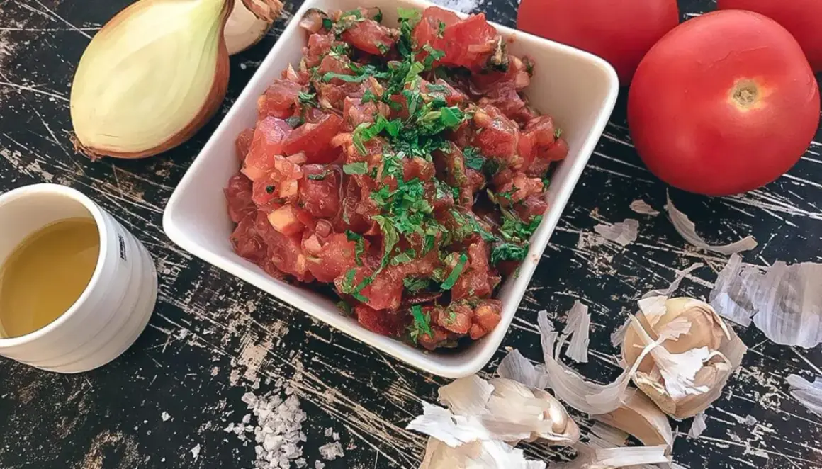 En skål med färsk tomatsalsa, redo för ett tomatsalsa recept. Ingredienser som lök, vitlök och tomater syns runt omkring.