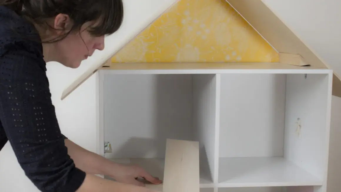 Une femme assemble une maison de poupée. Elle pose une pièce en bois dans une étagère blanche, sous un toit jaune à motifs.