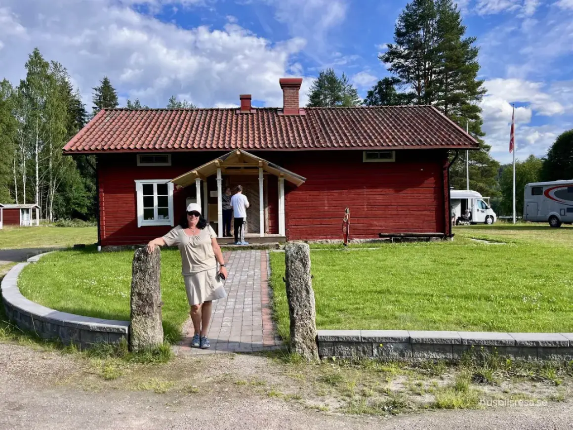 Värnäs camping: En kvinna står vid en röd stuga med tegeltak. Husbilar syns i bakgrunden under en blå himmel med moln.
