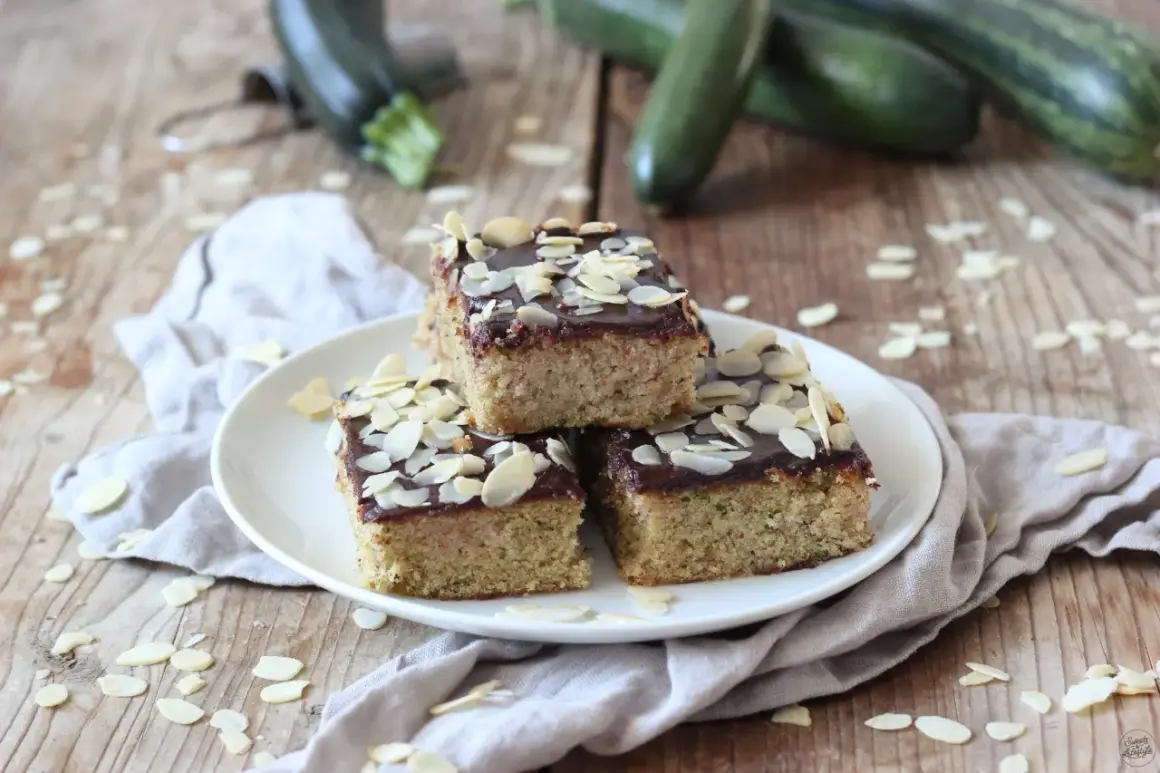 Saftiger Zucchini Kuchen mit Schokoladenglasur und Mandelblättchen, garniert mit frischen Zucchini im Hintergrund.