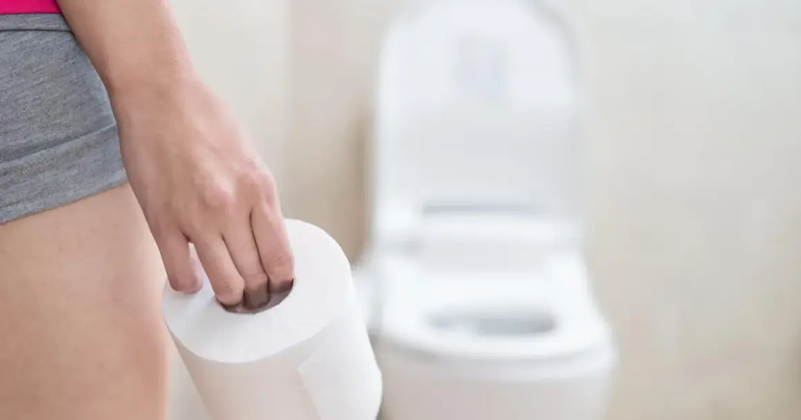Dłoń trzymająca rolkę papieru toaletowego przed toaletą. Może to sugerować problemy z wypróżnianiem, co powoduje zaparcia.