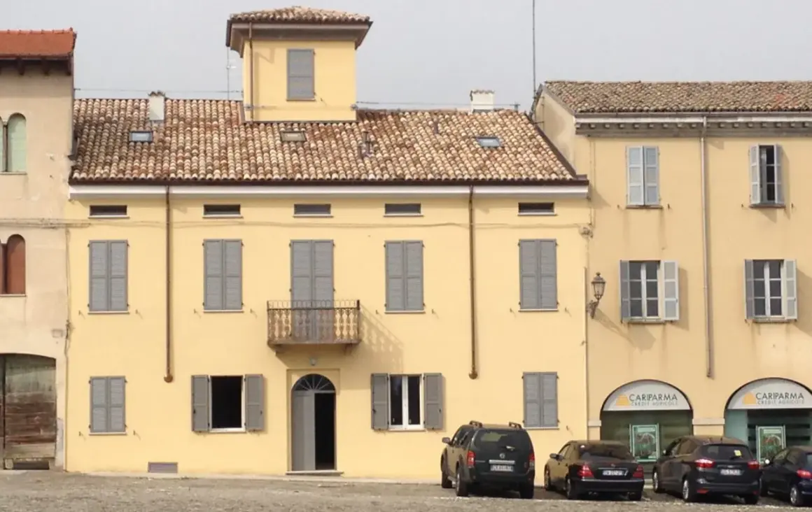 Edificio giallo con intonachino silossanico, tegole rosse, persiane grigie e balcone. Auto parcheggiate davanti.