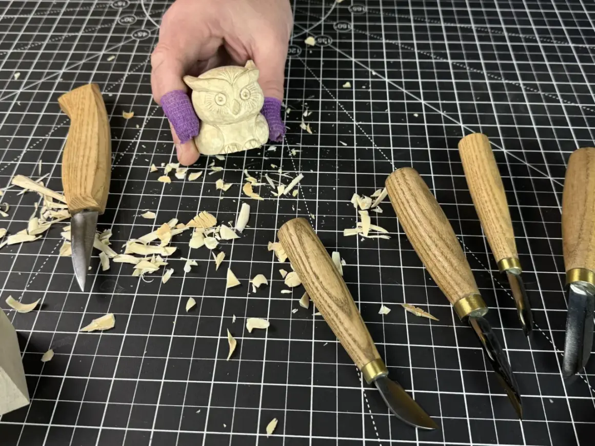 Une main tient une petite chouette en bois sculpté, entourée d'outils et de copeaux. Idéal pour une **débutant sculpture bois facile**.