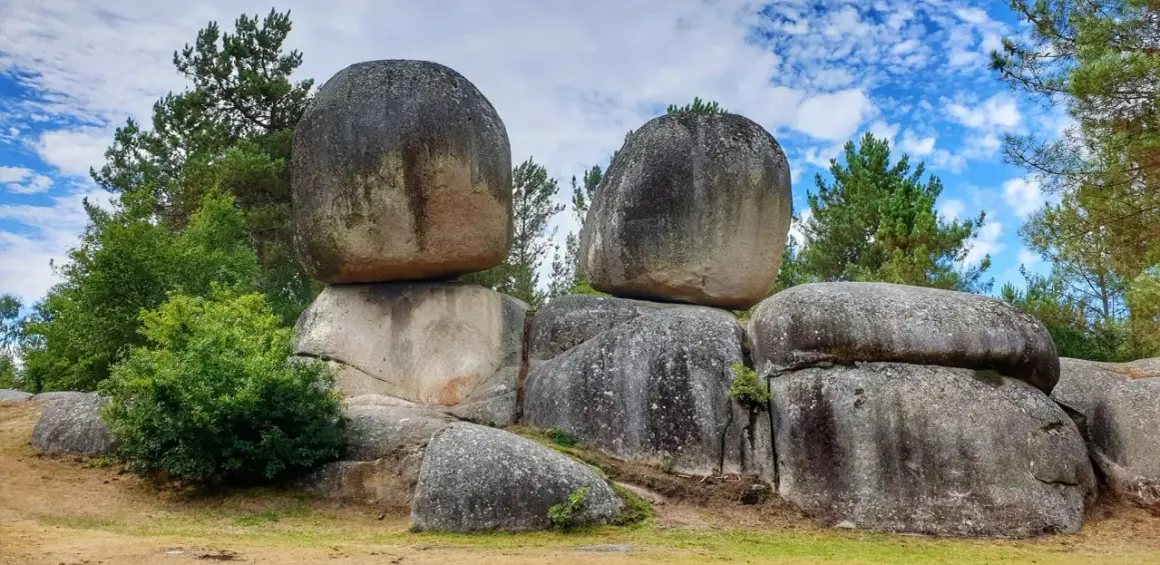Gigantescas rocas apiladas, como penas de rodas, desafían la gravedad en un paisaje boscoso bajo un cielo azul con nubes.