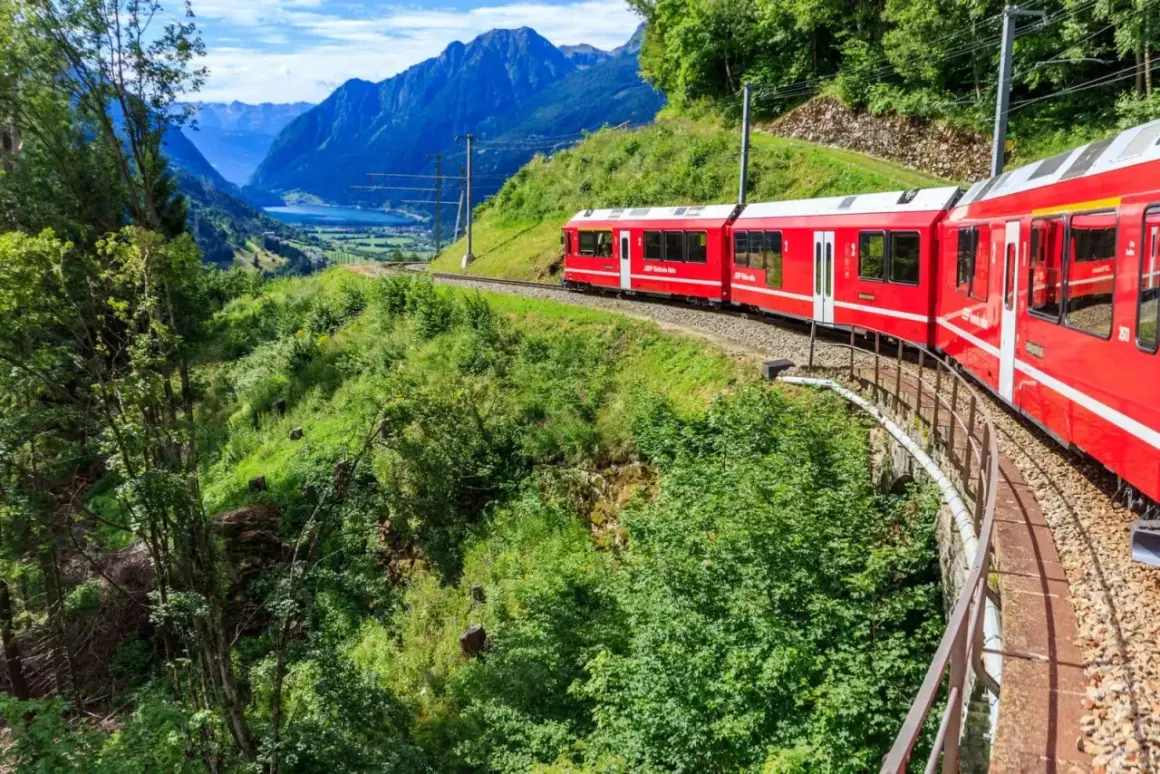 Röda tåget Bernina Express slingrar sig genom ett grönt alplandskap med en sjö och berg i bakgrunden. Undrar du över bernina express pris?