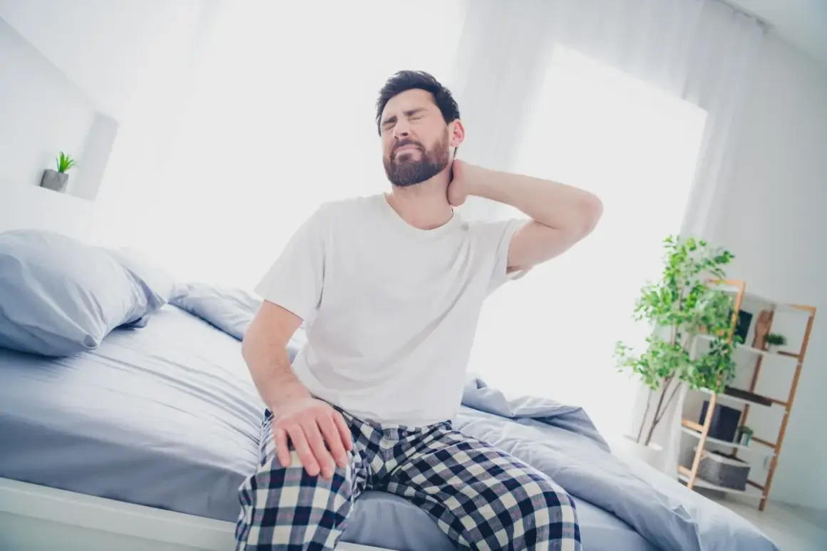 Uomo con torcicollo dopo aver dormito, si massaggia il collo con espressione sofferente, seduto sul letto.