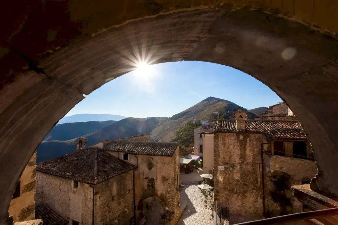 Solstrålar bryter fram genom en stenbåge över en pittoresk by i Abruzzo, Italien. Kulliga berg i bakgrunden.