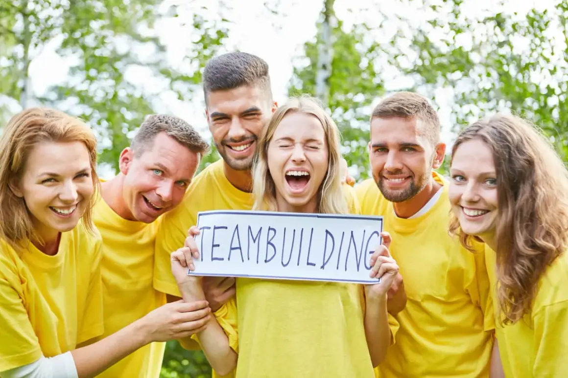 En grupp glada människor i gula tröjor håller upp en skylt med "TEAMBUILDING". De verkar ha det roligt, som om de precis svarat på några snabba roliga frågor.