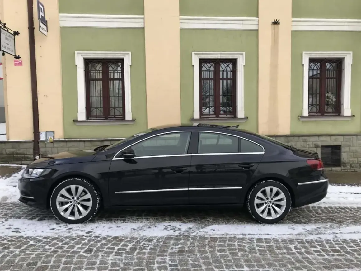 Elegancki, czarny Volkswagen Passat CC na zimowych oponach stoi na ośnieżonej, brukowanej ulicy przed kamienicą z zielonymi oknami.