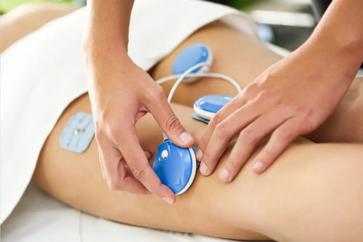 Fizykoterapia co to? Elektrostymulacja mięśni na udzie pacjenta. Terapeuta umieszcza elektrody.