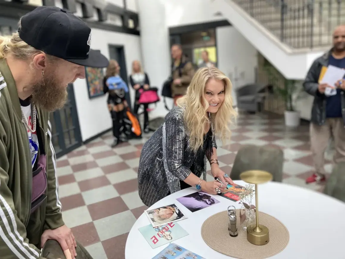 Ankie Bagger signerar bilder vid ett bord. En man med skägg och keps står bredvid.
