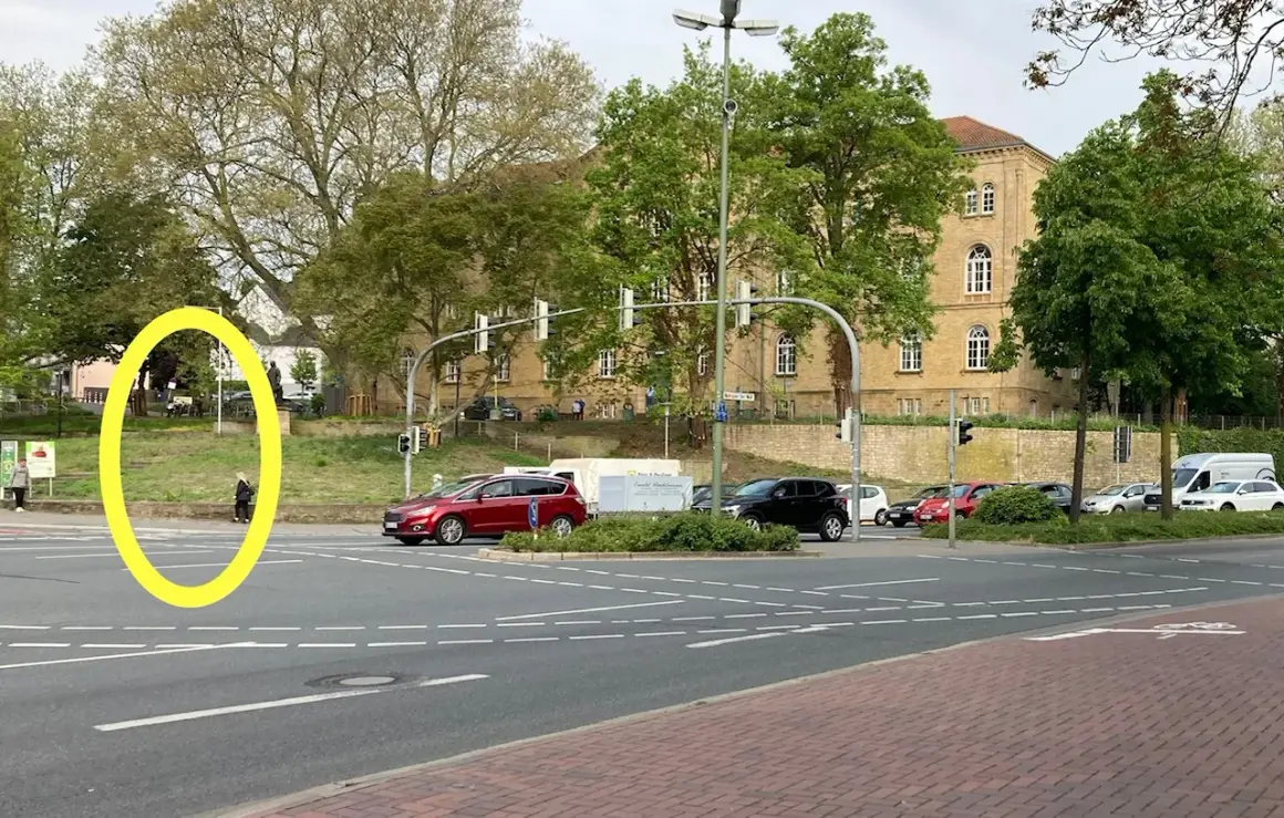 Eine Person wartet an der Ampel, um links abzubiegen. Autos stehen bereit, während im Hintergrund ein Gebäude und Bäume zu sehen sind.