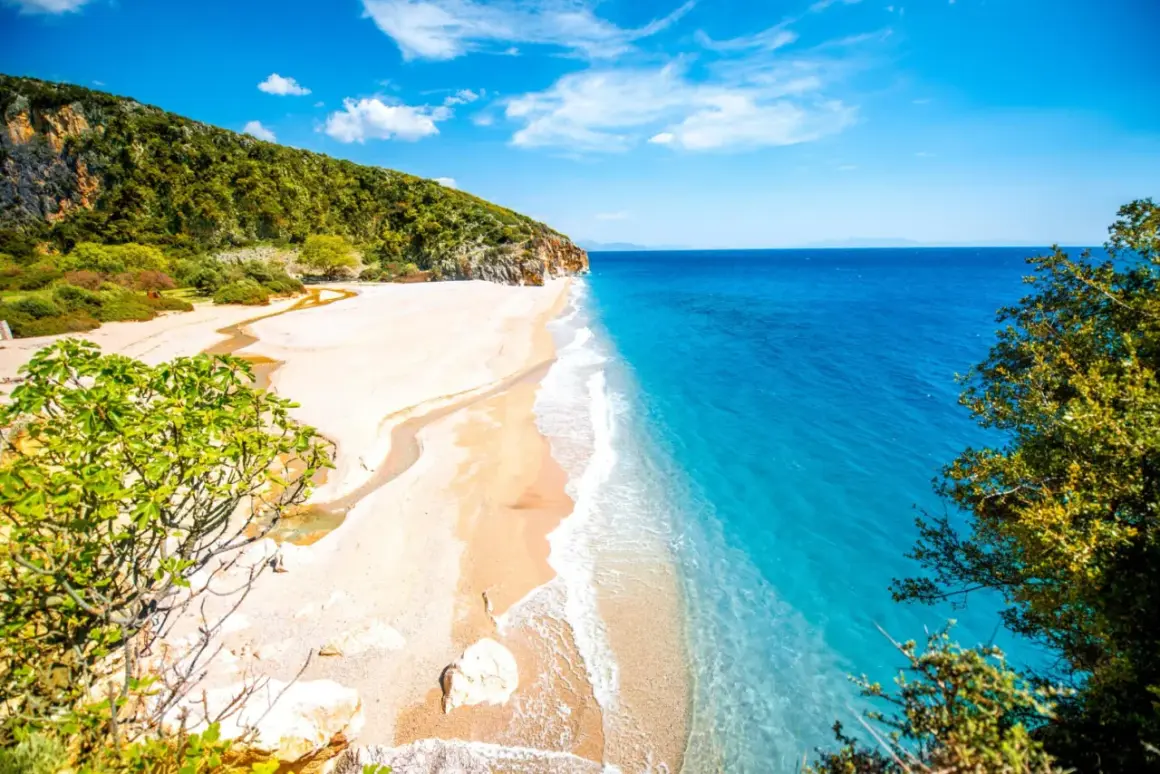 En orörd albanien strand med turkost vatten, vit sand och gröna kullar. En liten flod rinner ut i havet.
