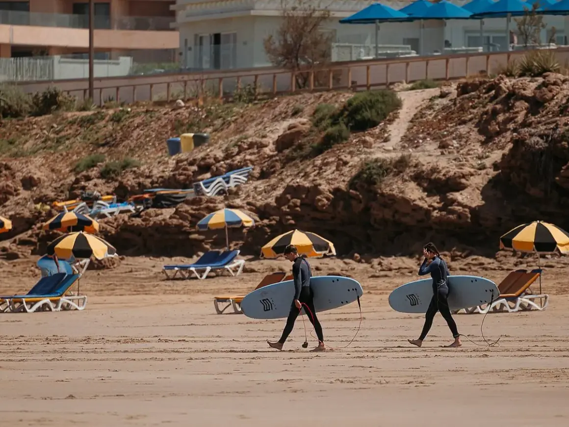 Deux surfeurs marchent sur la plage de Tamraght, Maroc, avec leurs planches sous le bras. Des parasols et des chaises longues sont installés sur le sable.