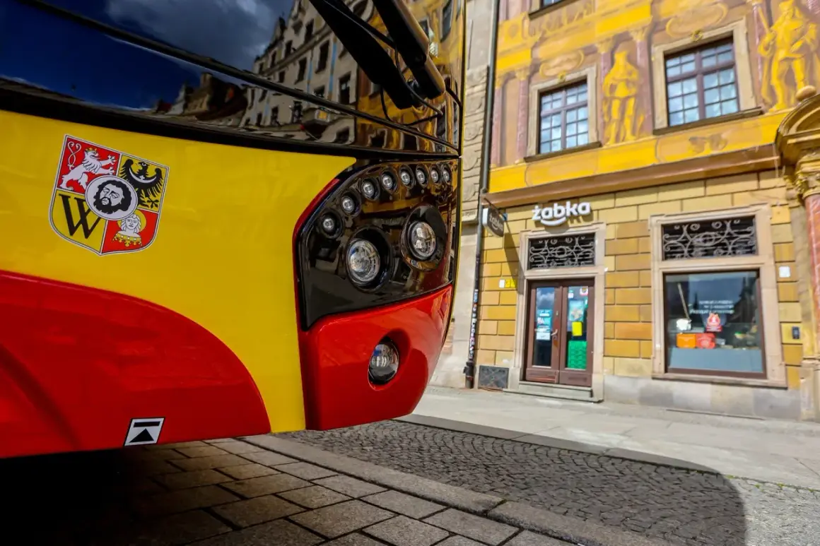 Żółto-czerwony autobus 143 Kraków z herbem miasta na błotniku, zaparkowany przed kamienicą z malowidłami i sklepem Żabka.