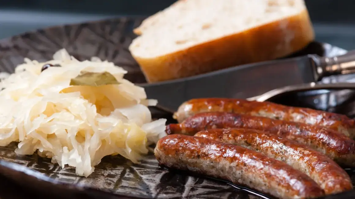 Herzhaftes Sauerkraut mit vier goldbraunen Würsten und einem Stück Brot auf einem dunklen Teller.