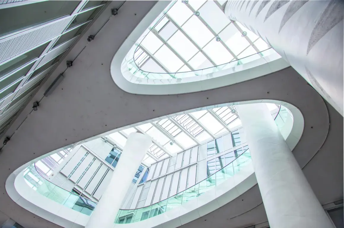 Nowoczesne wnętrze z białymi kolumnami i szklanymi balustradami, przez które wpada obfite światło dzienne.