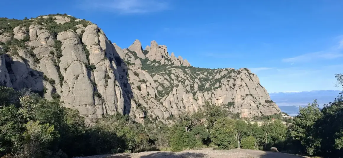 Imponentes rocas de Montserrat bajo un cielo azul, con vegetación verde y la creu de Sant Miquel en la distancia.