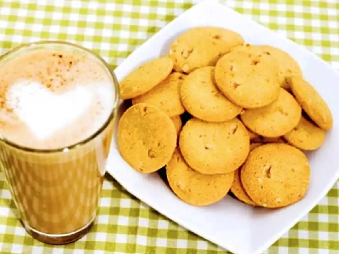 A glass of coffee with a heart-shaped foam art next to a plate of golden brown, round cookies.
