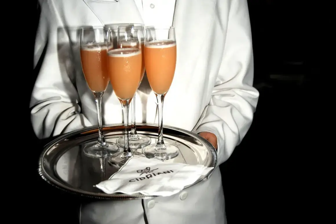 Kellner serviert Bellinis auf einem Silbertablett. So trinkt man Bellini: ein erfrischender Cocktail aus Prosecco und Pfirsichpüree.