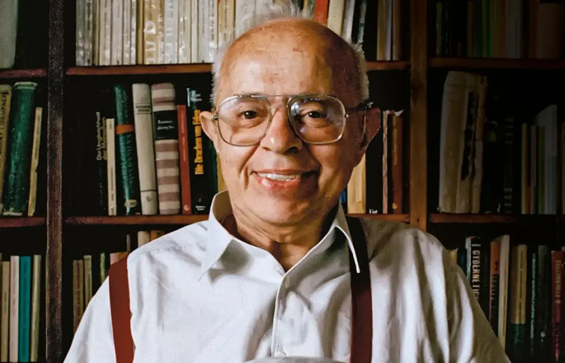 Uśmiechnięty Stanisław Lem, autor powieści Solaris, siedzi przed regałami pełnymi książek.