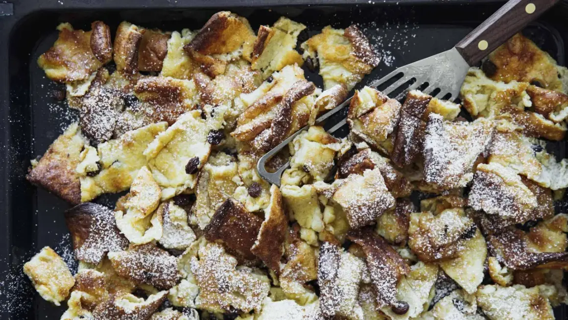 Kaiserschmarrn aus dem Backofen auf einem Blech, mit Puderzucker bestäubt und Rosinen. Ein Gabel-Schaber liegt bereit.