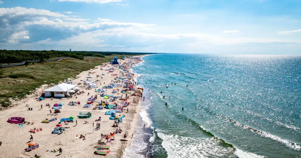 Tłum ludzi na plaży w Darłowie, korzystających z uroków Bałtyku. Wiele kolorowych parawanów i namiotów, a w wodzie kąpiący się. To typowe atrakcje Darłowa latem.