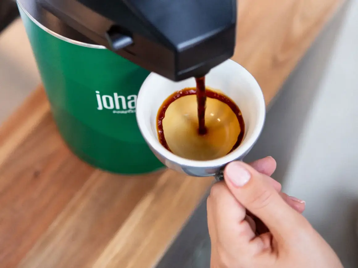 En hand håller en kopp medan kaffe rinner ner från en maskin. Frågan om hur många matskedar kaffe per kopp är relevant för att få den perfekta smaken.