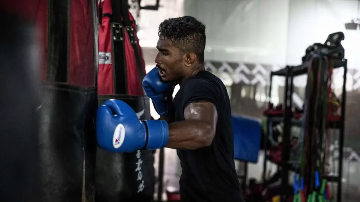Boxer in blue gloves throwing a powerful cross punch at a heavy bag, sweat glistening on his arms.