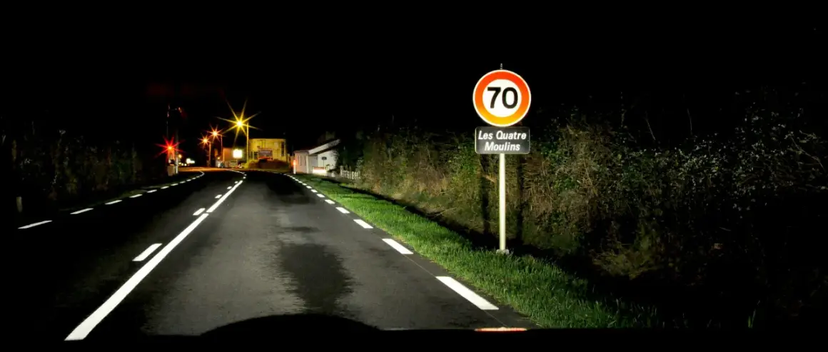 Route de nuit avec marquage au sol blanc lumineux. Panneau "Les Quatre Moulins" et limite de vitesse à 70.