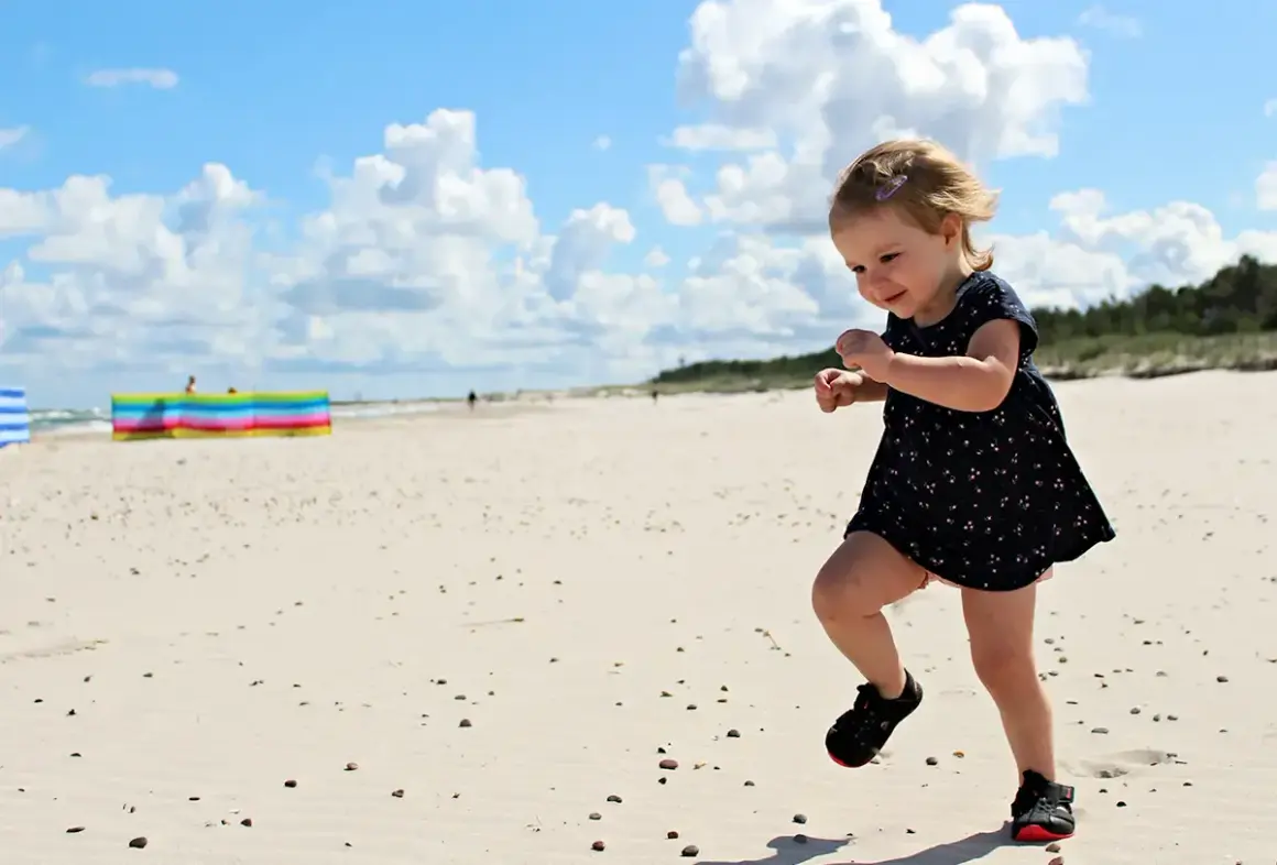 Radosna wycieczka dla dzieci na plażę. Mała dziewczynka w letniej sukience biega po piasku, ciesząc się słońcem i chmurami.