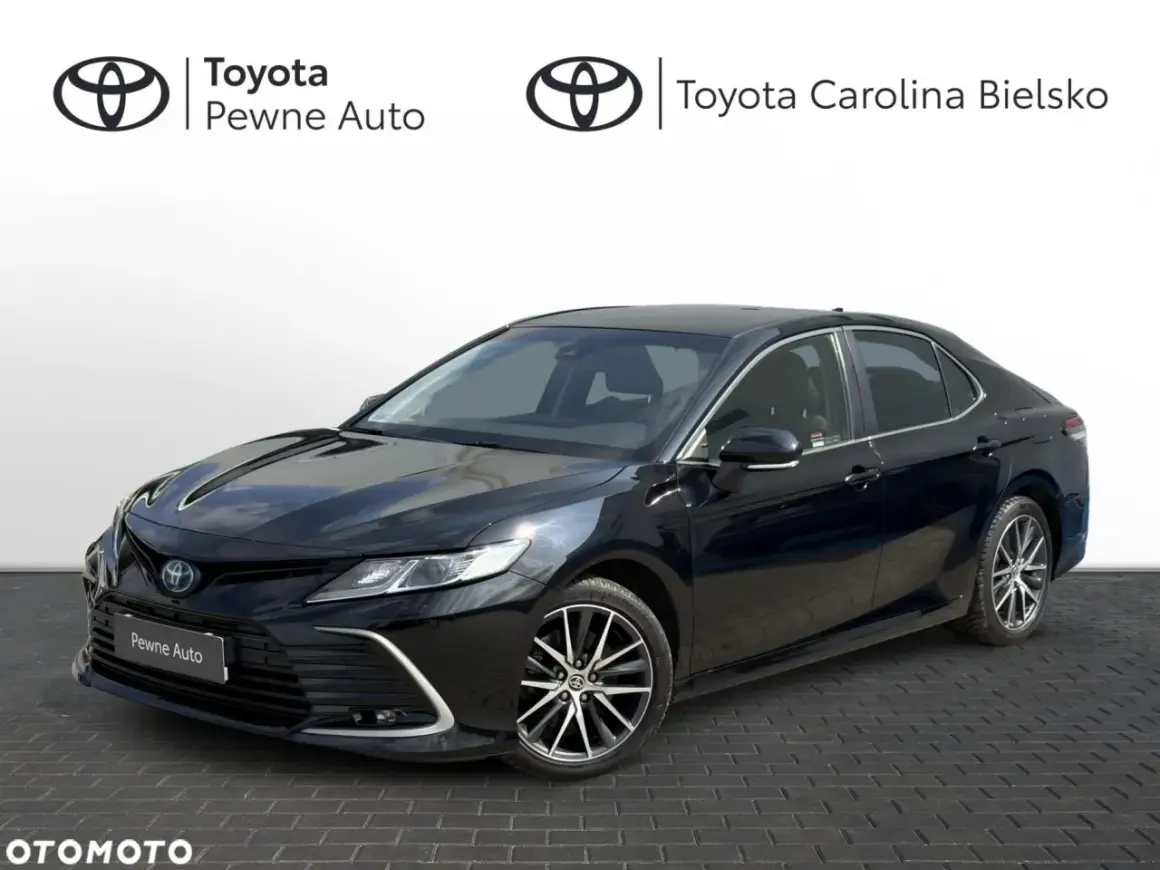 Elegancka, czarna Toyota Camry z napisem "Pewne Auto" na grillu. Lśniący lakier odbija światło, podkreślając dynamiczną sylwetkę sedana.
