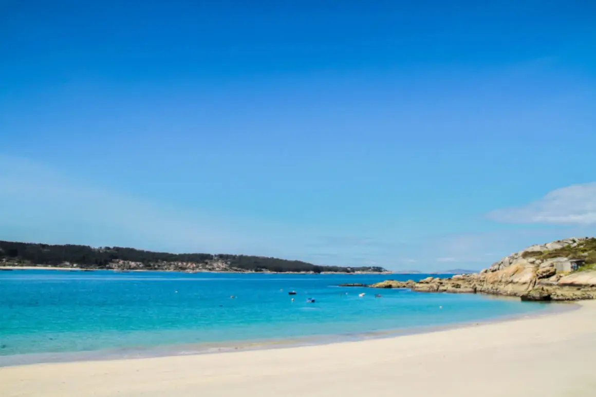 Arena blanca y aguas turquesas en la playa de Menduiña, con barcas a lo lejos y una costa rocosa.