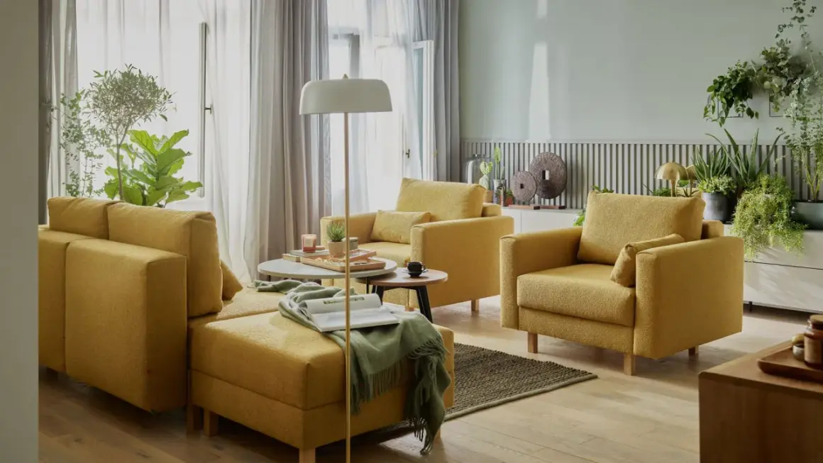 Przytulny salon bez telewizora. Żółte meble wypoczynkowe, stoliki kawowe, lampa podłogowa i mnóstwo zielonych roślin tworzą relaksującą atmosferę.