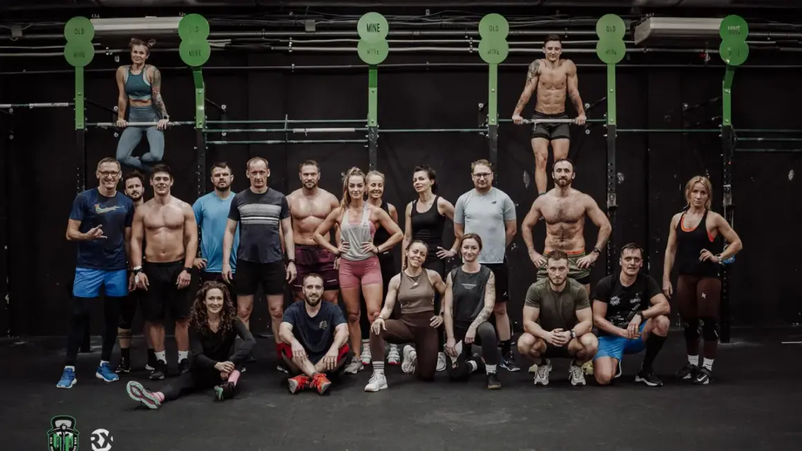 Grupa osób w strojach sportowych pozuje na tle sali treningowej CrossFit.