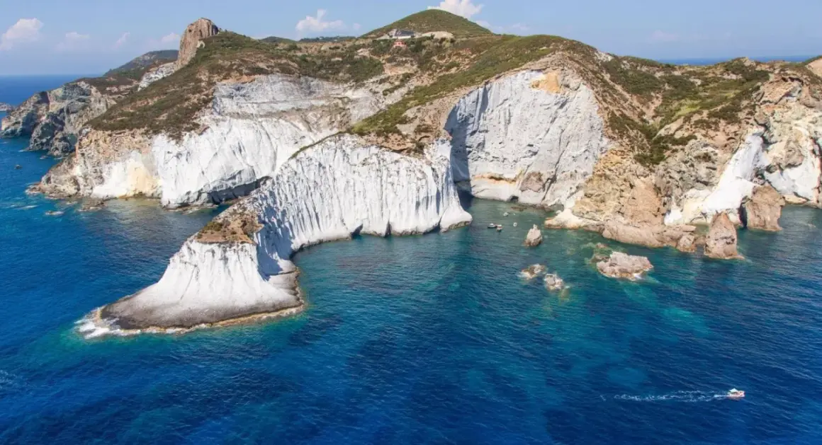 Scogliere bianche a picco sul mare blu intenso di Ponza. Un piccolo gozzo solca le acque cristalline, mentre la natura scolpisce la costa.