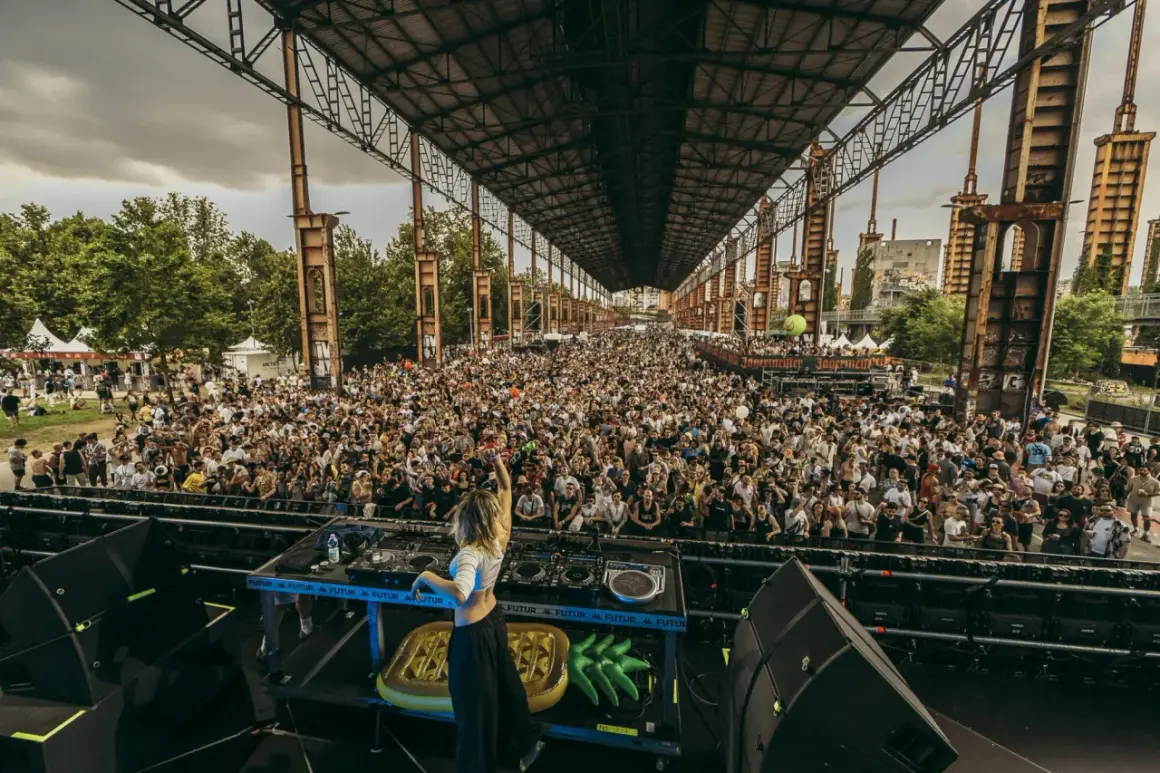 DJ sul palco, folla immensa sotto una struttura industriale, un'atmosfera da festival con il logo "Kappa" visibile.