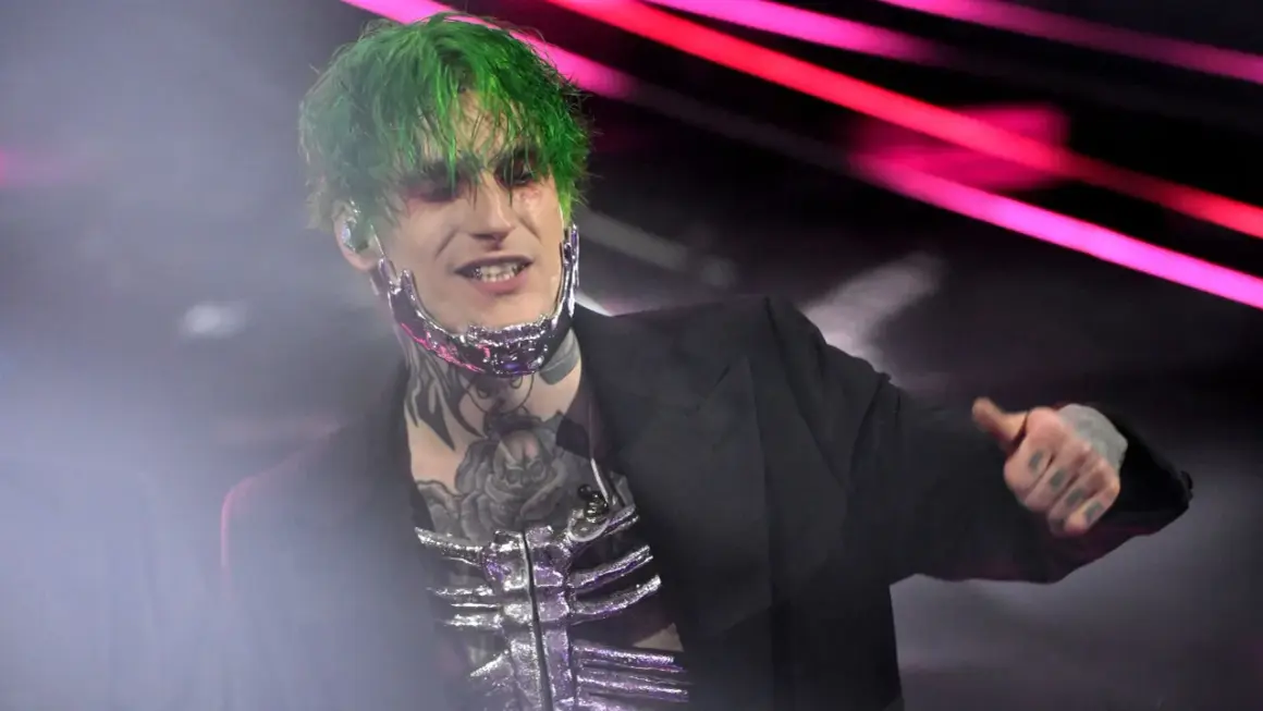 Theo la sad, con capelli verdi e trucco drammatico, indossa un abito nero e una maschera metallica.